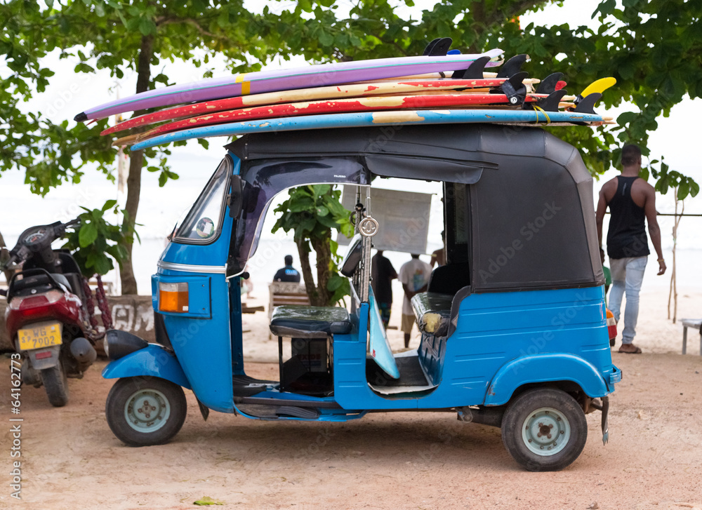 Blue tuk tuk vehicle transporting surfboards on the roof.