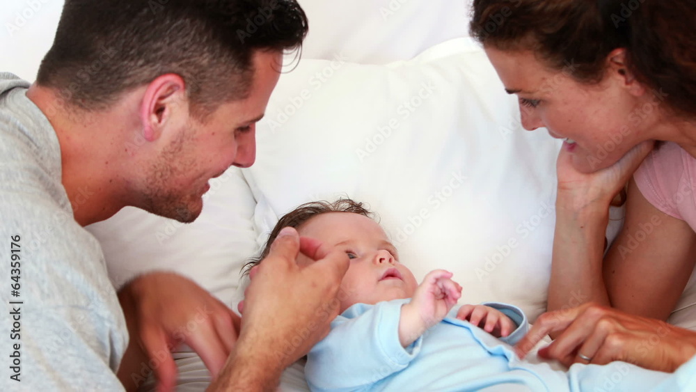 Baby boy in blue babygro being tickled by parents on bed Baby boy in blue babygro being tickled by parents on bed