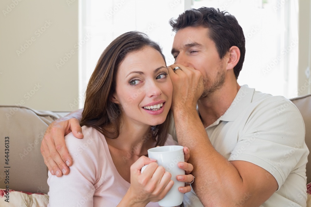 Man whispering secret into a happy womans ear in living room Man whispering secret into a happy womans ear in living room