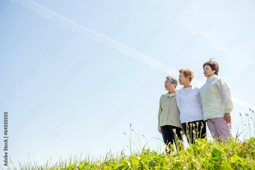 草原の丘の上に立って空を見上げる高齢の３人の日本人女性