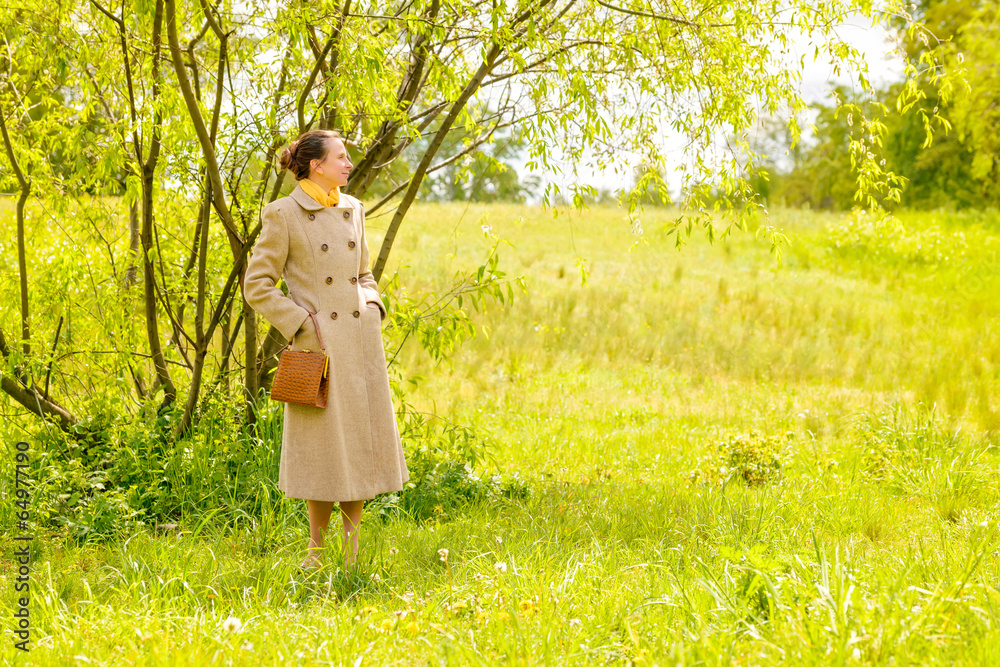 An elegant senior businesswoman, with a coat and a yellow scarf, waiting in the park under the spring sun An elegant senior businesswoman, with a coat and a yellow scarf, waiting in the park under the spring sun