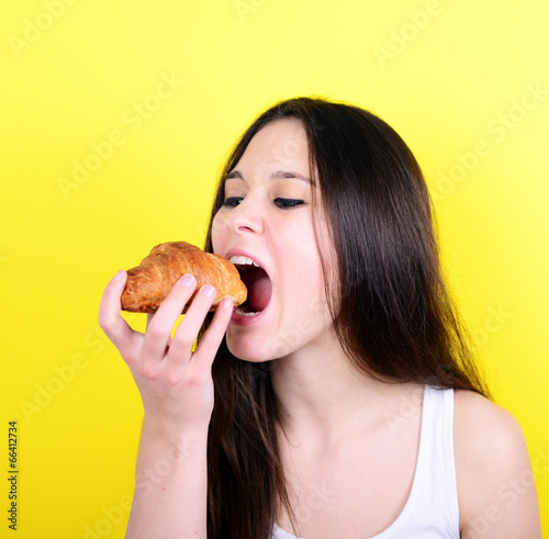 Portrait of young female eating croissant against yellow backgro