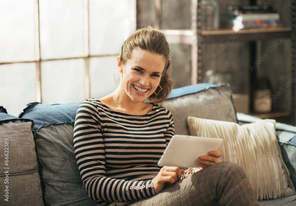 Obraz premium Smiling young woman sitting on sofa and using tablet pc