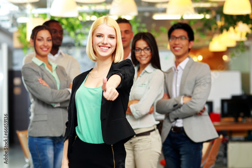 Businesswoman giving thumb up to camera in front of colleagues