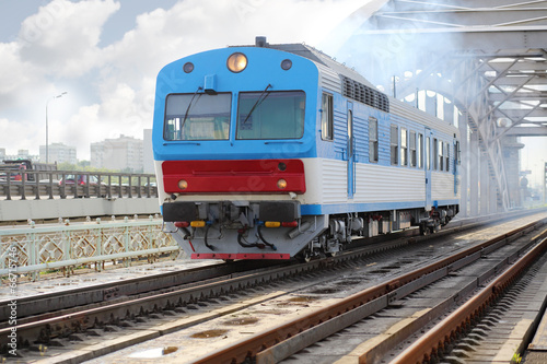Small blue train rides rails on railway bridge at summer day.