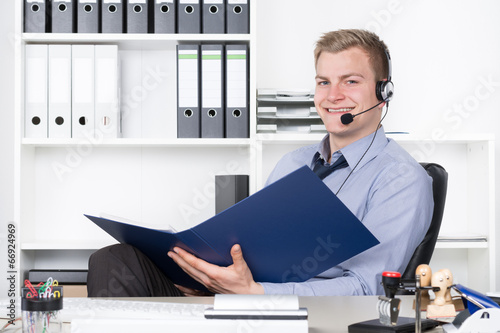 Junger lächelnder Mann mit Headset und Aktenordner im Büro