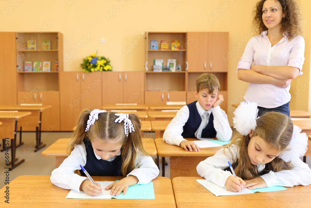 Obraz premium Two girls and boy write at school desks in classroom and teacher