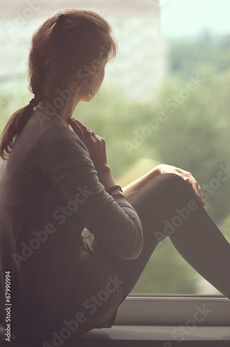 Girl sitting in the shadows on the window sill looking out the w