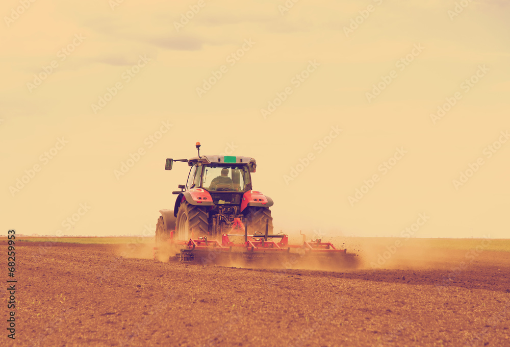 Naklejka premium Red tractor driven by farmer cultivating land at spring