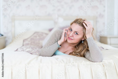 Natural cheerful blonde lying on bed and phoning in bright bedro
