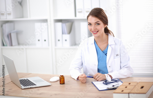 Beautiful young smiling female doctor sitting at the desk and