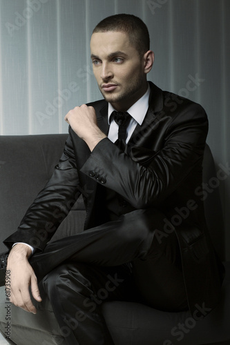 elegant young fashion man in tuxedo sitting on a sofa