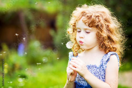Beautiful little curly girl blowing dandelion, horizontal shot