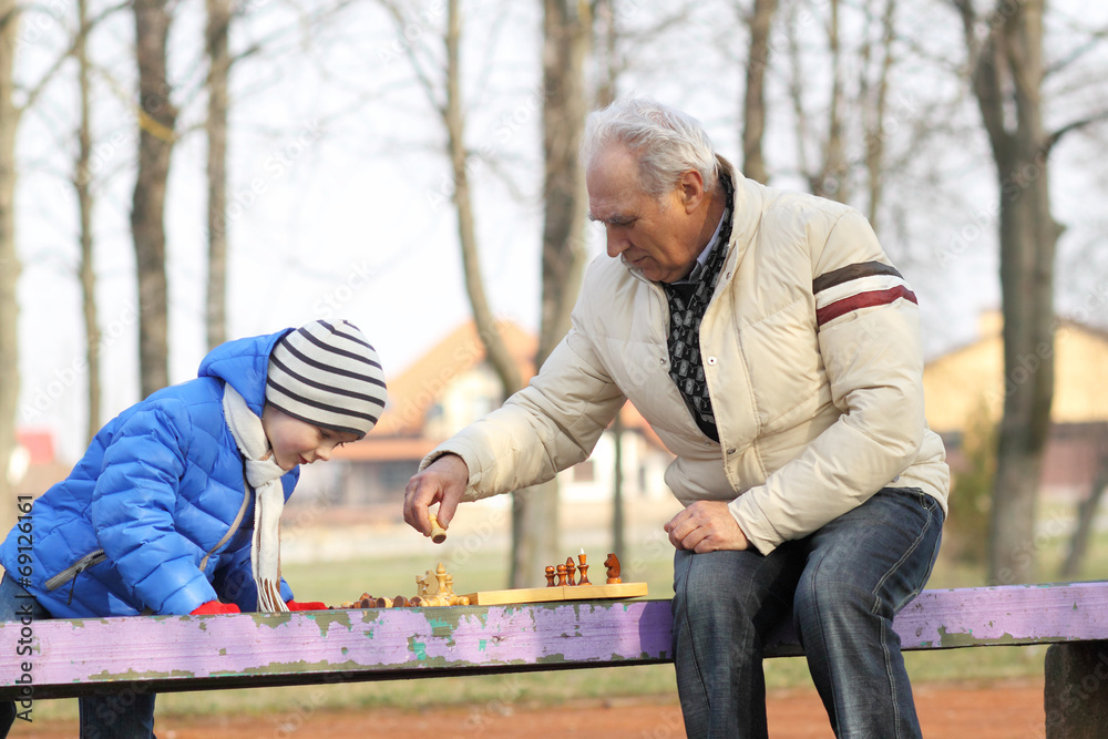 Fototapeta premium Grandfather and grandson playing chess on a bench outdoors