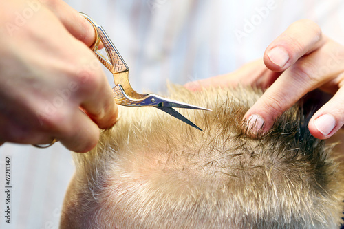 Hairdresser trimming blonde hair of young boy by scissors