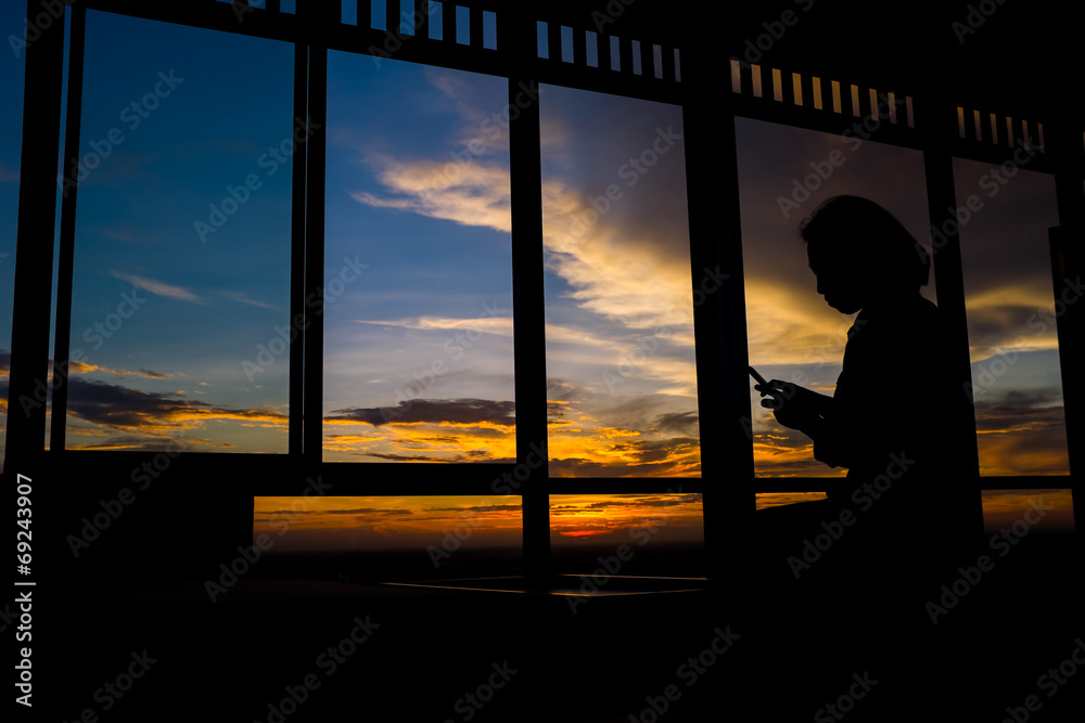 Girl playing smart phone by window at sunset with twilight sky