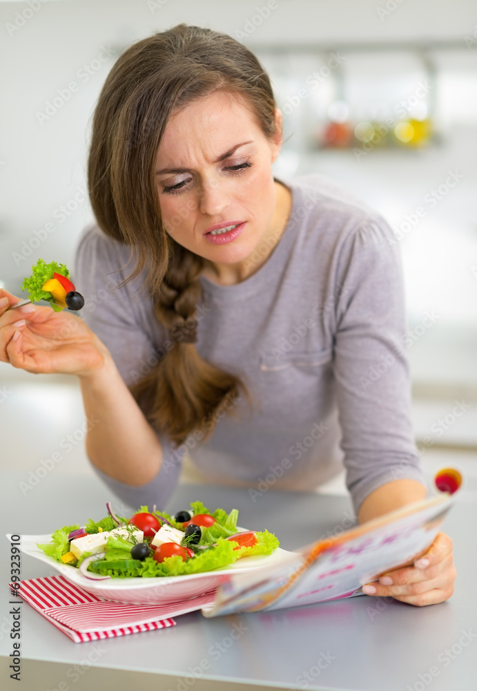 Young woman eating greek salad in kitchen and reading magazine