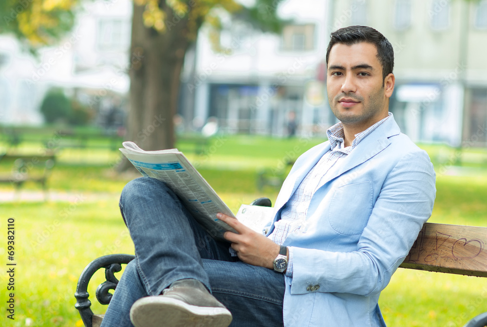 Obraz premium Young businessman sitting on bench and reading newspaper