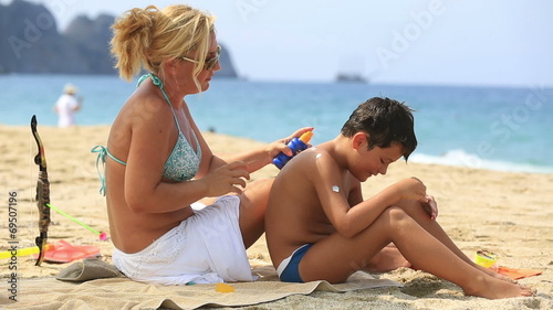 mother applying sunscreen to son on the beach at summertime