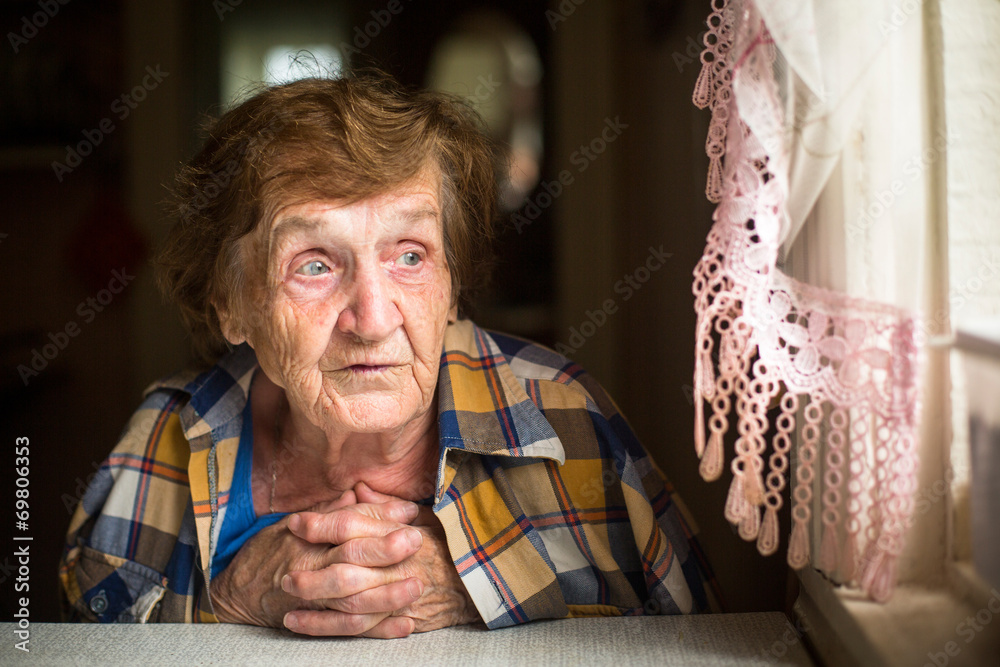 Old woman sitting at a table near the window in his house. - obrazy, fototapety, plakaty 