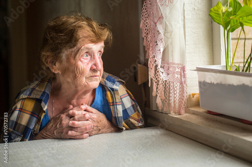 Old lonely woman sitting near the window in his house. photo