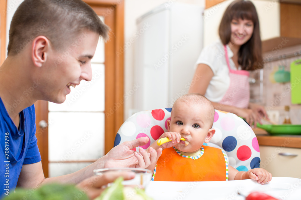 father feeding his baby and mother cooking at kitchen father feeding his baby and mother cooking at kitchen