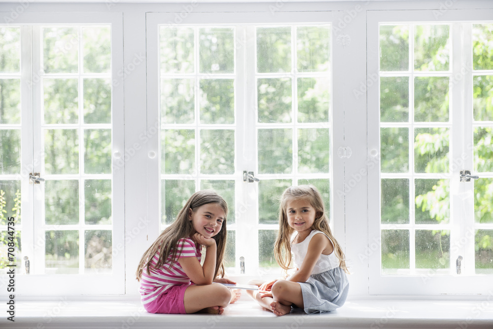 Two girls with digital tablet sitting by large window Two girls with digital tablet sitting by large window
