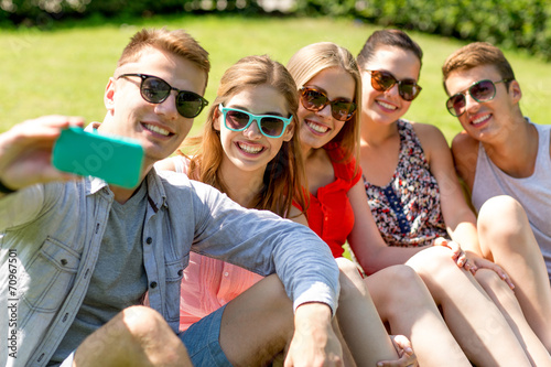 smiling friends with smartphone sitting on grass
