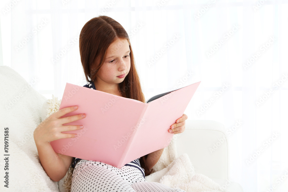 Beautiful little girl reading book on sofa in room Beautiful little girl reading book on sofa in room