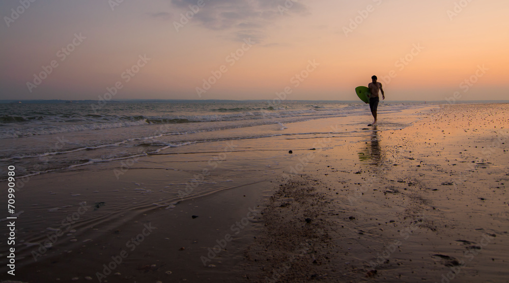 A single surfer on the shore, with the sun setting behind him