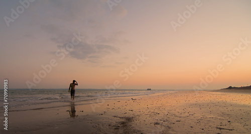 A single surfer on the shore, with the sun setting behind him