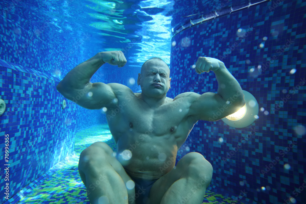 Foto Stock Bodybuilder in swimming trunks sits on bottom of pool underwater  | Adobe Stock