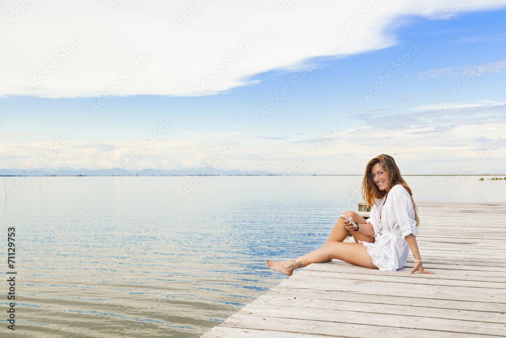 Smiling beautiful young woman sitting on a pier and using a mobi
