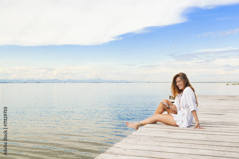 Smiling beautiful young woman sitting on a pier and using a mobi Smiling beautiful young woman sitting on a pier and using a mobi