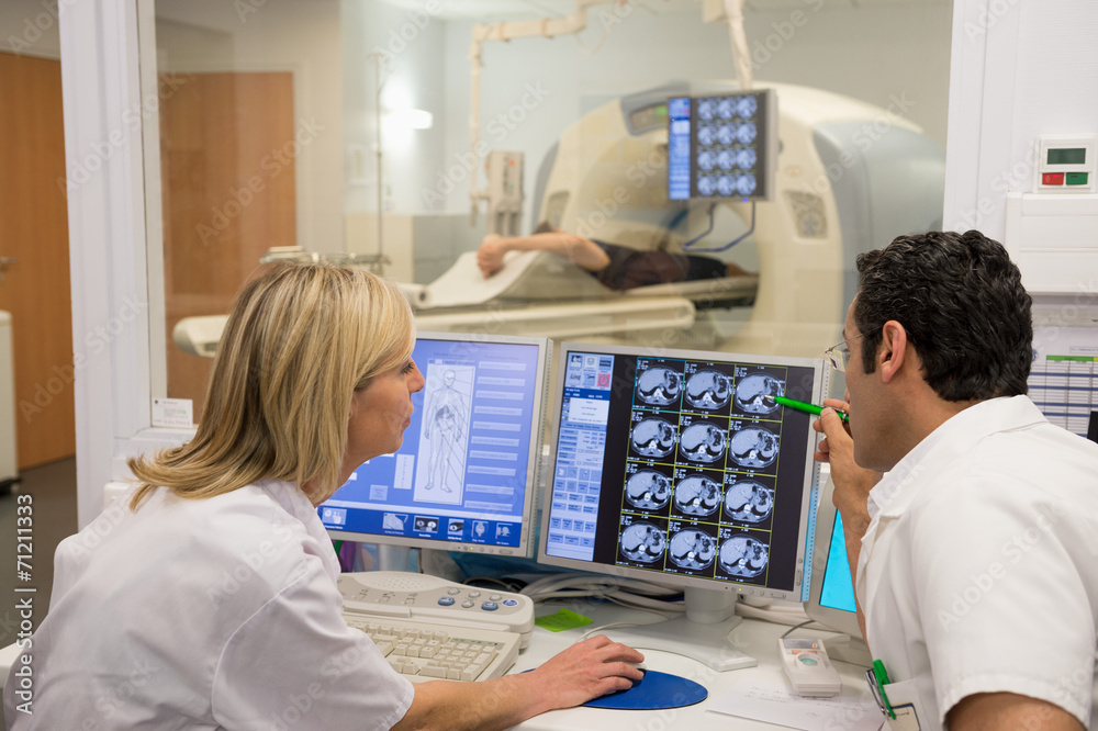 Doctors examining scan on computer with patient on MRI scanner in background Doctors examining scan on computer with patient on MRI scanner in background