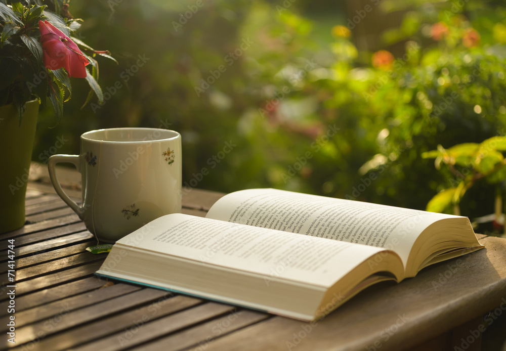 Obraz premium Cup of tea and open book on wooden table in garden.