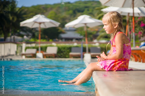 Adorable girl seat at side of swimming pool with legs splasing a
