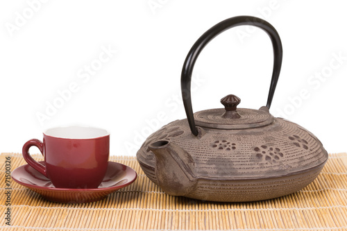 Iron teapot with red cup of tea on wooden mat.