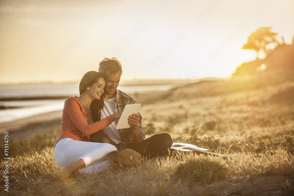 handsome couple sitting on the beach at sunset and using a digit