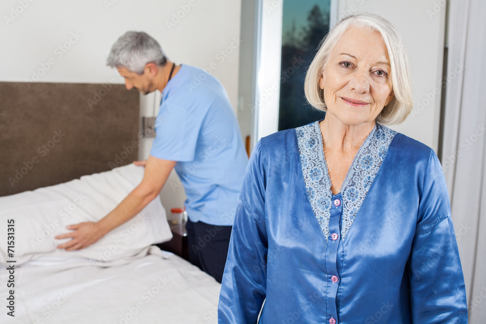 Senior Woman With Caretaker Making Bed At Nursing Home