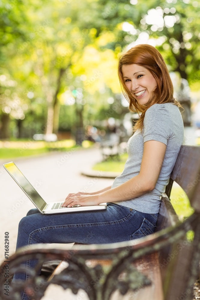 Redhead sitting on bench using laptop smiling at camera