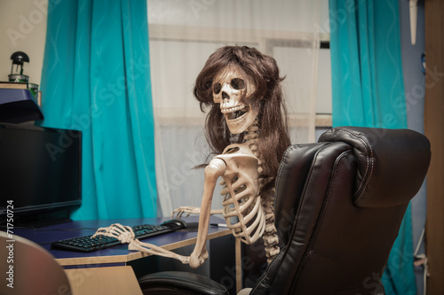 smiling skeleton on black leather chair behind the desktop