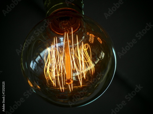 Single edison light bulb with the filaments glowing orange against a dark black Fototapeta