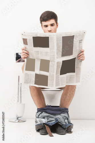 Young handsome man sitting on toilet and reading magazine.