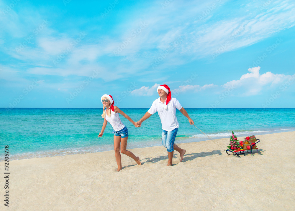Happy couple at beach in red santa hats with gifts, sled and dec