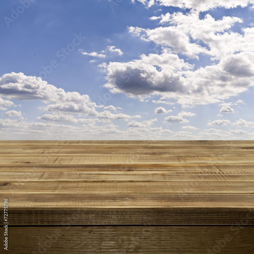 Empty wooden table top against a blue sky with fluffy white clou