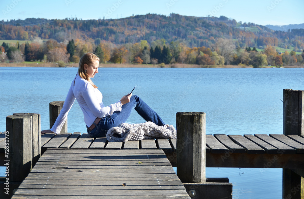 Girl reading from a tablet on the wooden jetty against a lake. S Girl reading from a tablet on the wooden jetty against a lake. S