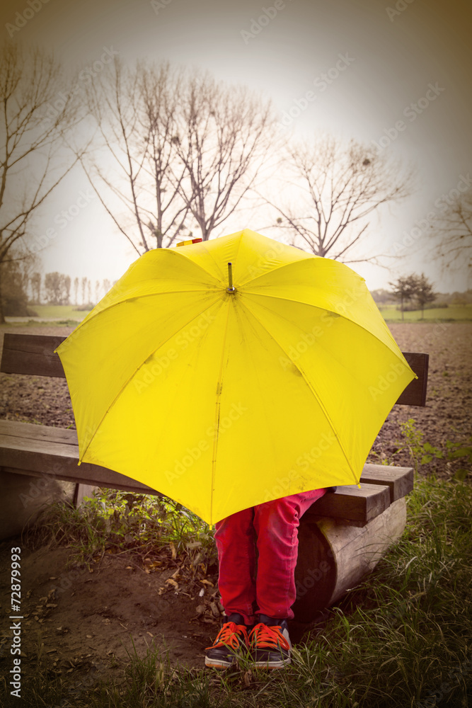 boy sitting on a bench hidden by a yellow umbrella