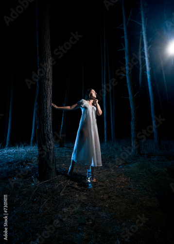 woman in white nightgown looking at ray of light at night forest