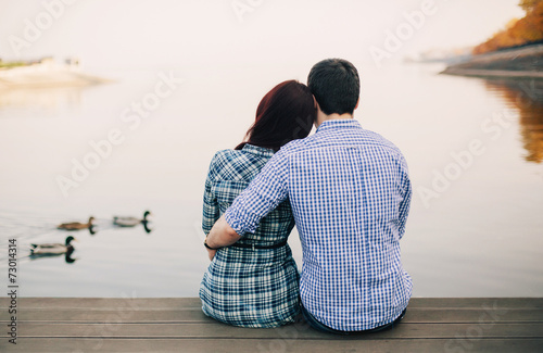 Rear view of a romantic young couple sitting on the river dock i